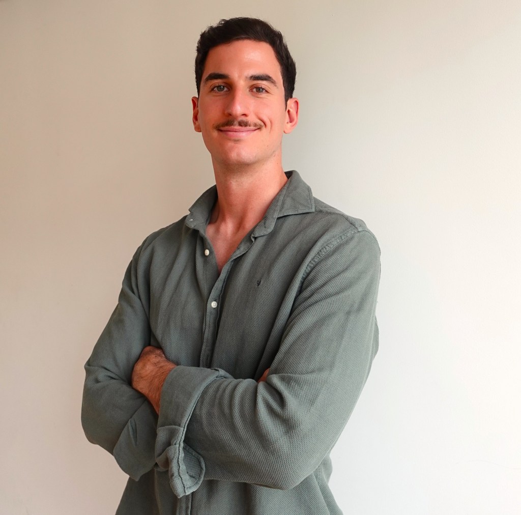 A young man with a mustache, wearing a green shirt, stands confidently with arms crossed against a light-colored background.